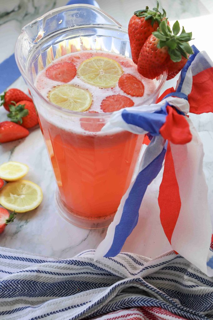 A pitcher of Strawberry Puree Lemonade with lemon and strawberry slices, garnished with whole strawberries and tied with red, white, and blue ribbon, sits on a marble surface near a cut lemon and strawberries.