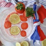 A clear pitcher filled with pink, fizzy Strawberry Puree Lemonade is garnished with lemon and strawberry slices. Whole strawberries and lemon wedges surround the pitcher, which is decorated with a red, white, and blue ribbon. A striped cloth is in the background.