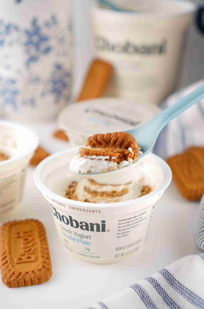 A spoon with a Biscoff cookie and Greek yogurt is held above an open container of Chobani Greek yogurt—a simple biscoff yogurt recipe surrounded by more cookies and yogurt containers in the background.