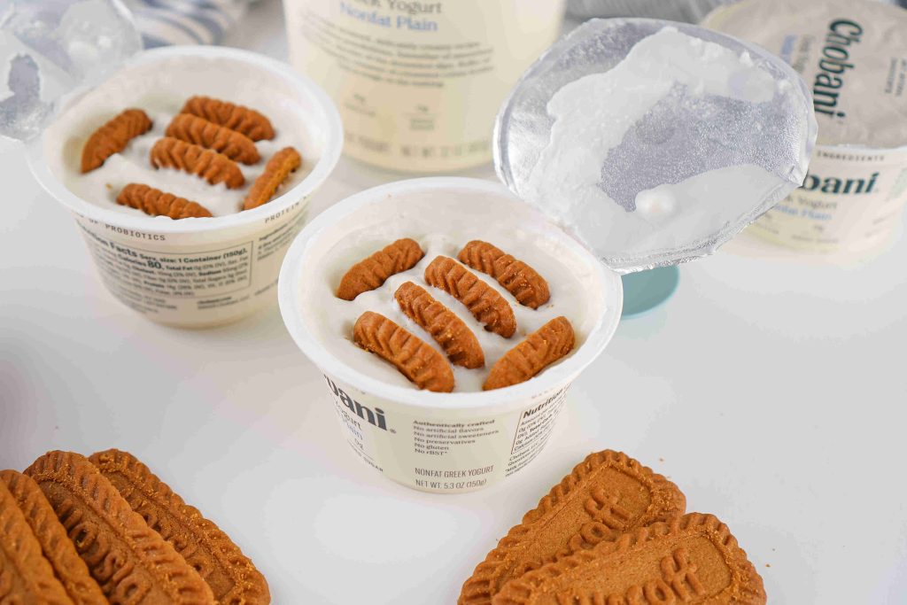A container of Chobani Greek yogurt topped with several rectangular cookies, resembling a biscoff yogurt recipe, with more yogurt containers and cookies nearby on a white surface.