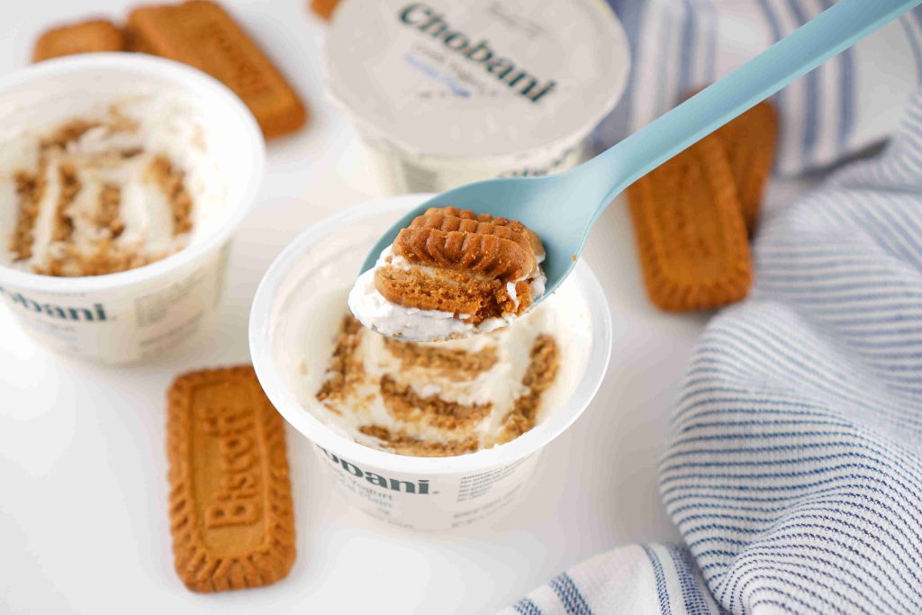 A blue spoon holds a bite of Greek yogurt layered with crumbled Biscoff cookies above an open Chobani yogurt cup, creating a tempting biscoff yogurt recipe scene, with extra cookies and cups nearby on a white surface next to a striped cloth.