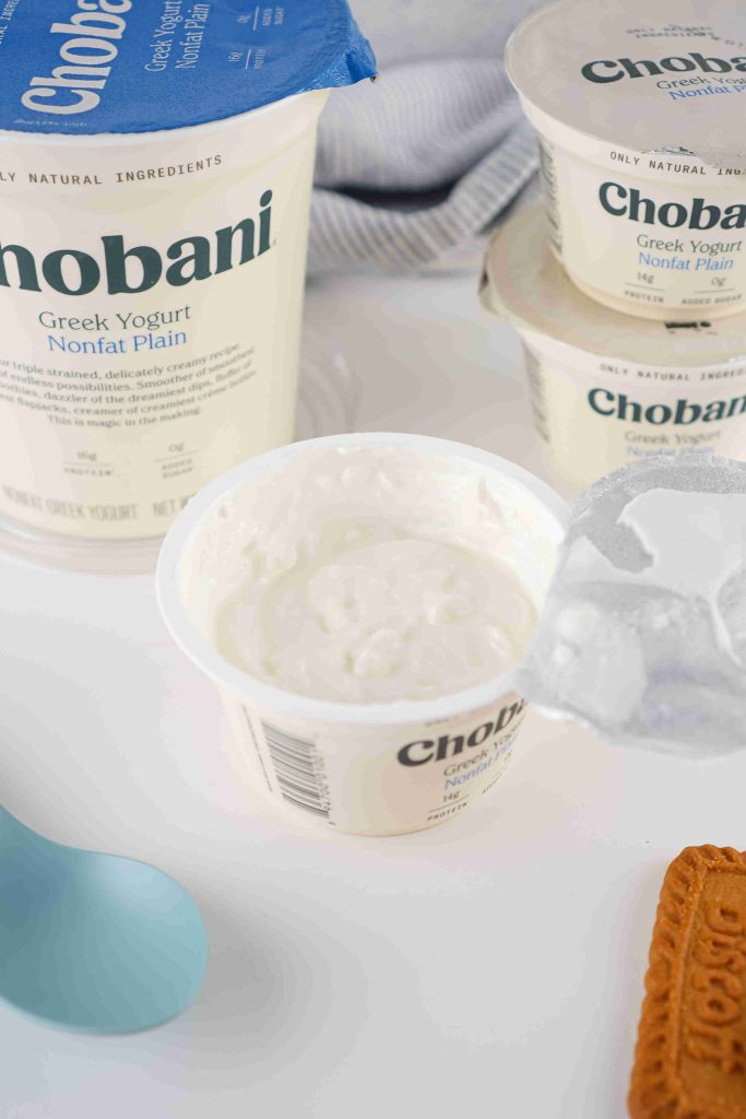 An opened container of Chobani Greek Yogurt Nonfat Plain sits on a white surface, its foil lid peeled back, ready to inspire your next biscoff yogurt recipe. Nearby are unopened yogurt containers and a blue spoon.