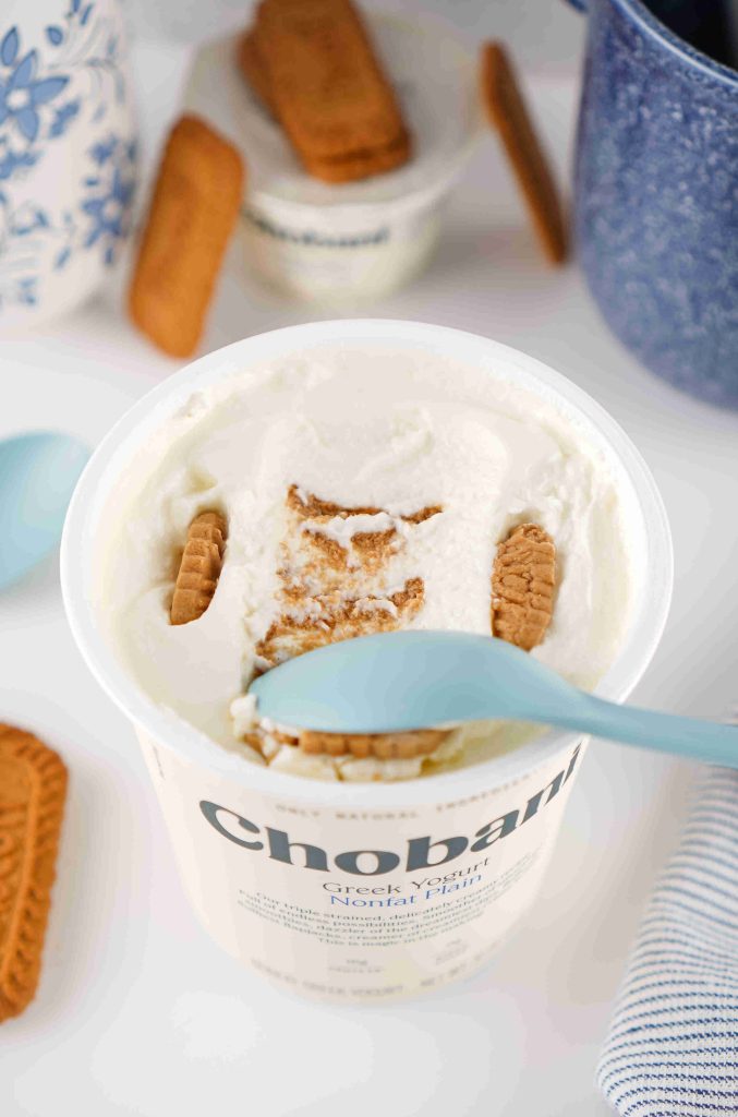 A container of Chobani Greek yogurt with crumbled cookie pieces on top, inspired by a biscoff yogurt recipe. A blue spoon rests inside the yogurt, and cookies are scattered around on a white surface.