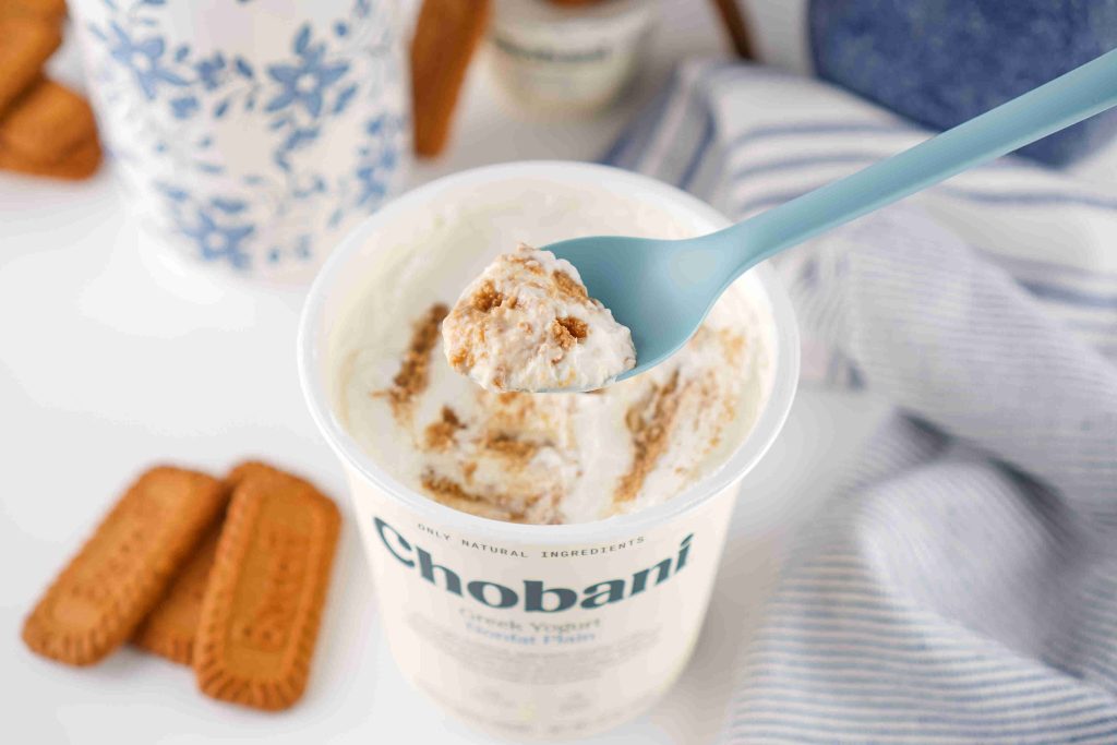 A blue spoon holds a scoop of Chobani Greek yogurt with visible cookie crumbs above an open container, hinting at a tasty biscoff yogurt recipe. Biscuit cookies and a blue patterned cup are in the background.