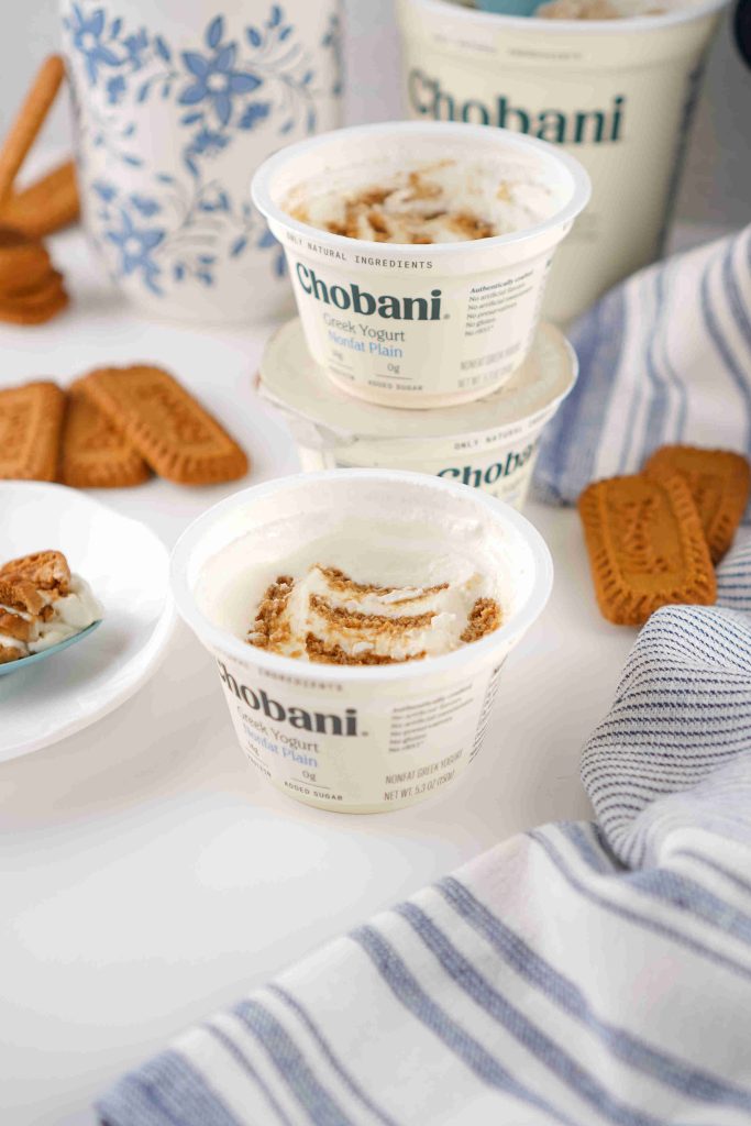 Three containers of Chobani Greek yogurt with crumbled cookies, inspired by a biscoff yogurt recipe, are stacked on a white surface, surrounded by whole cookies, a blue-and-white patterned vase, and a blue-striped towel.