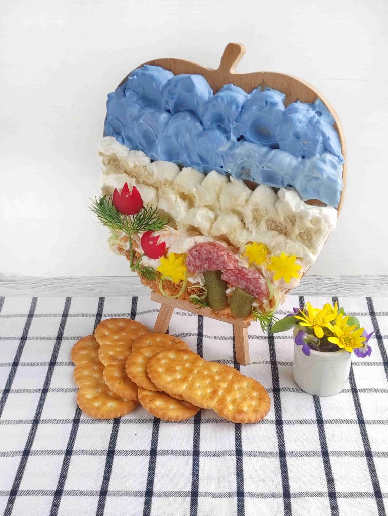 A charcuterie board shaped like an apple, designed as a delightful cheese canvas and decorated with colorful spreads, veggies, and salami, sits on a mini easel beside a stack of crackers and a small cup of yellow and purple flowers.