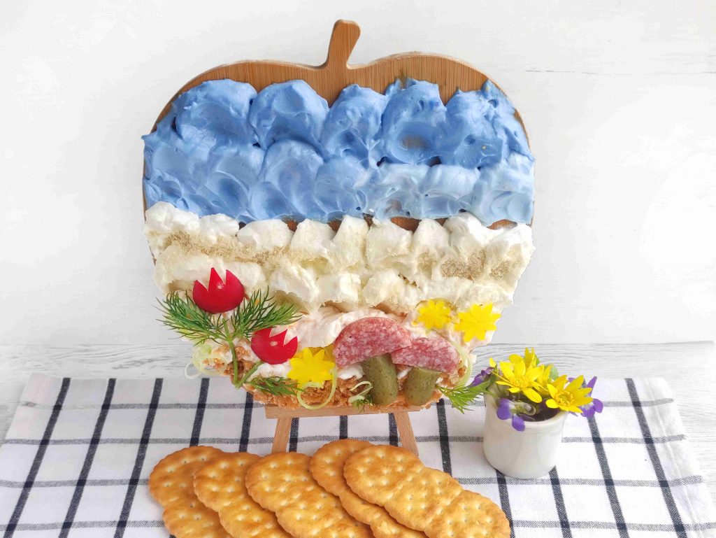 A cutting board with food arranged to resemble a landscape, using blue and white spreads as a cheese canvas, alongside bread, meats, herbs, and flowers. Crackers are in front and a small cup with more flowers sits on a checkered cloth.