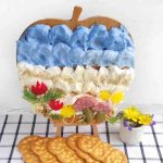 A cutting board with food arranged to resemble a landscape, using blue and white spreads as a cheese canvas, alongside bread, meats, herbs, and flowers. Crackers are in front and a small cup with more flowers sits on a checkered cloth.