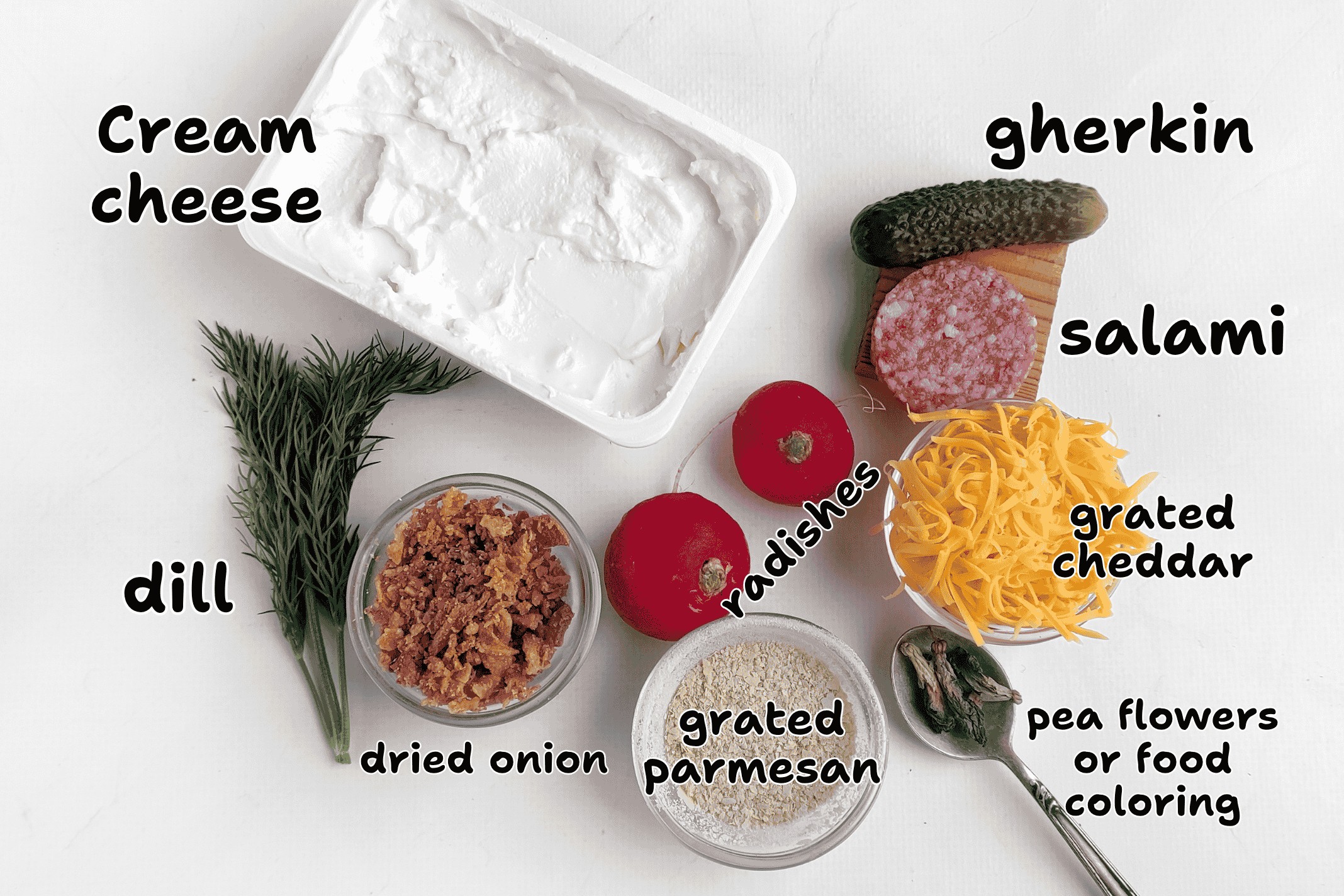 Various ingredients including cream cheese, dill, dried onion, grated parmesan, radishes, gherkin, salami, grated cheddar, and a spoon with pea flowers or food coloring are displayed on a white surface—ready to become your ultimate cheese canvas for culinary creativity.