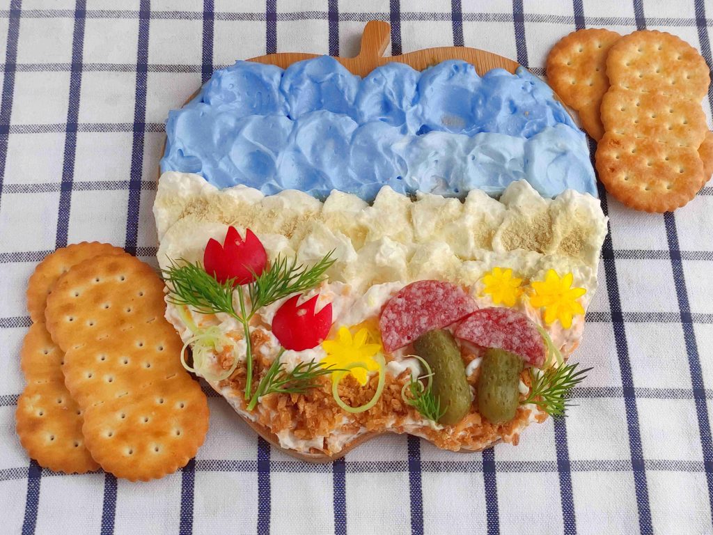Decorative appetizer cake shaped like Ukraine with blue and white layers, garnished with herbs, pickles, meat, crackers, and edible flowers—all beautifully arranged on a cheese canvas—placed atop a checked cloth with extra crackers on the side.