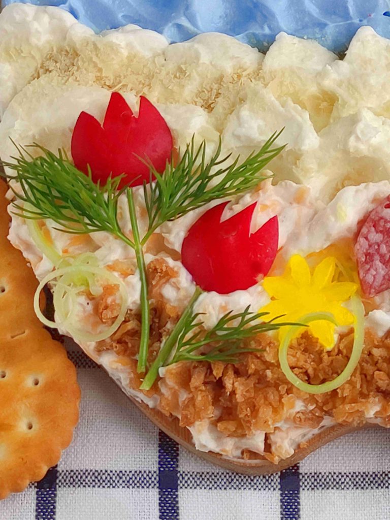 A colorful salad topped with whipped cream, tomato tulip flowers, dill, yellow pepper, crumbs, and two crackers on a checkered cloth creates a delightful cheese canvas for your next gathering.