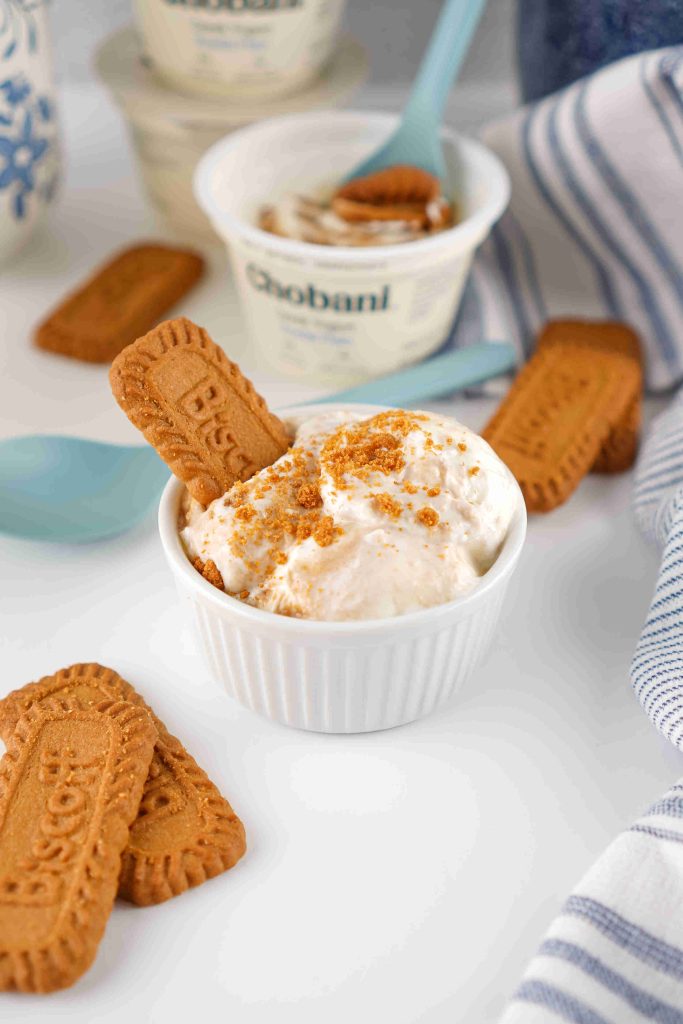 A small white bowl of creamy Biscoff yogurt recipe dessert topped with crushed Biscoff cookies and a whole cookie, surrounded by more cookies, a cup of Chobani yogurt, blue utensils, and a striped towel on a white surface.