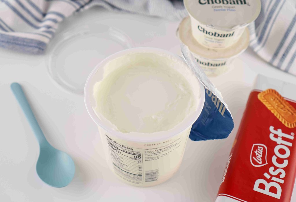 An open container of Chobani Greek yogurt sits on a white surface next to a blue spoon, a pack of Lotus Biscoff cookies, and two smaller sealed Chobani cups—perfect inspiration for your next biscoff yogurt recipe. A striped towel is in the background.
