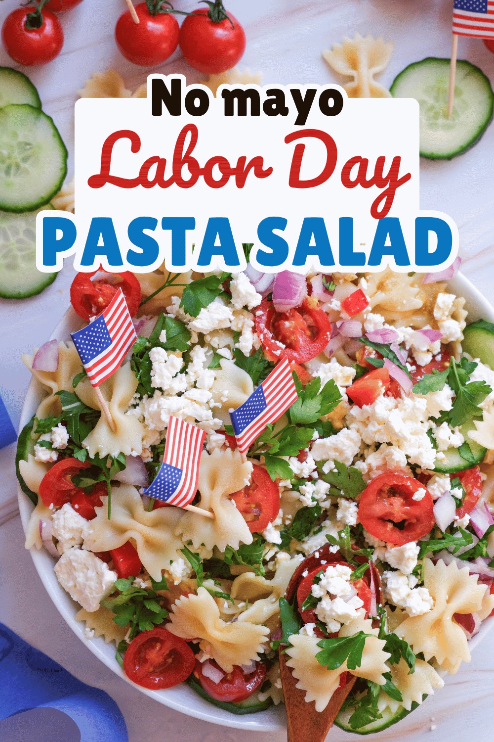A bowl of pasta salad with tomatoes, feta cheese, parsley, and American flag toothpicks. Sliced cucumber and utensils are nearby. The text reads: No mayo labor day pasta salad recipe.