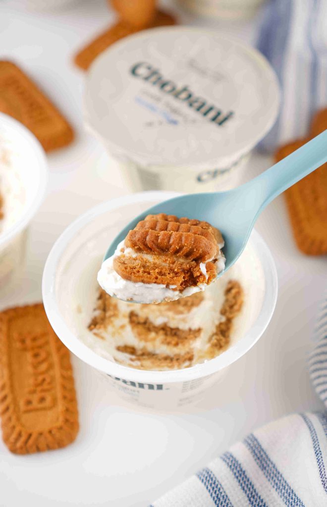 A spoon holds a piece of Biscoff cookie and creamy yogurt above an open Chobani cup—perfect inspiration for your next Biscoff yogurt recipe, with whole cookies and other yogurt cups in the background.