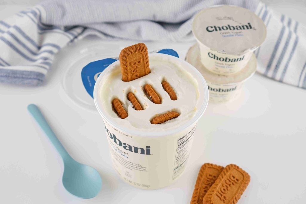 A container of Chobani Greek yogurt is topped with several Biscoff cookies, creating a tempting biscoff yogurt recipe. More cookies, unopened yogurt cups, a blue spoon, and a striped cloth are also visible.