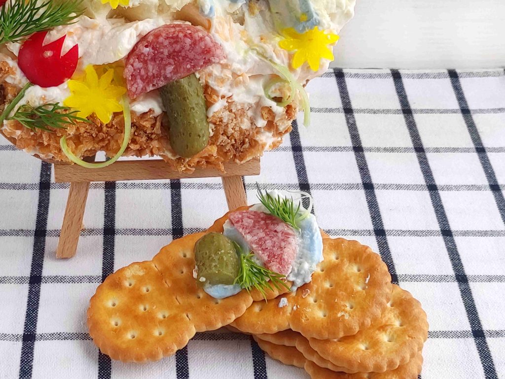 Crackers topped with cream spread, a slice of salami, a small pickle, dill, and a piece of lettuce are artfully arranged on a checkered cloth, with a decorated serving displayed like a cheese canvas on a mini easel in the background.