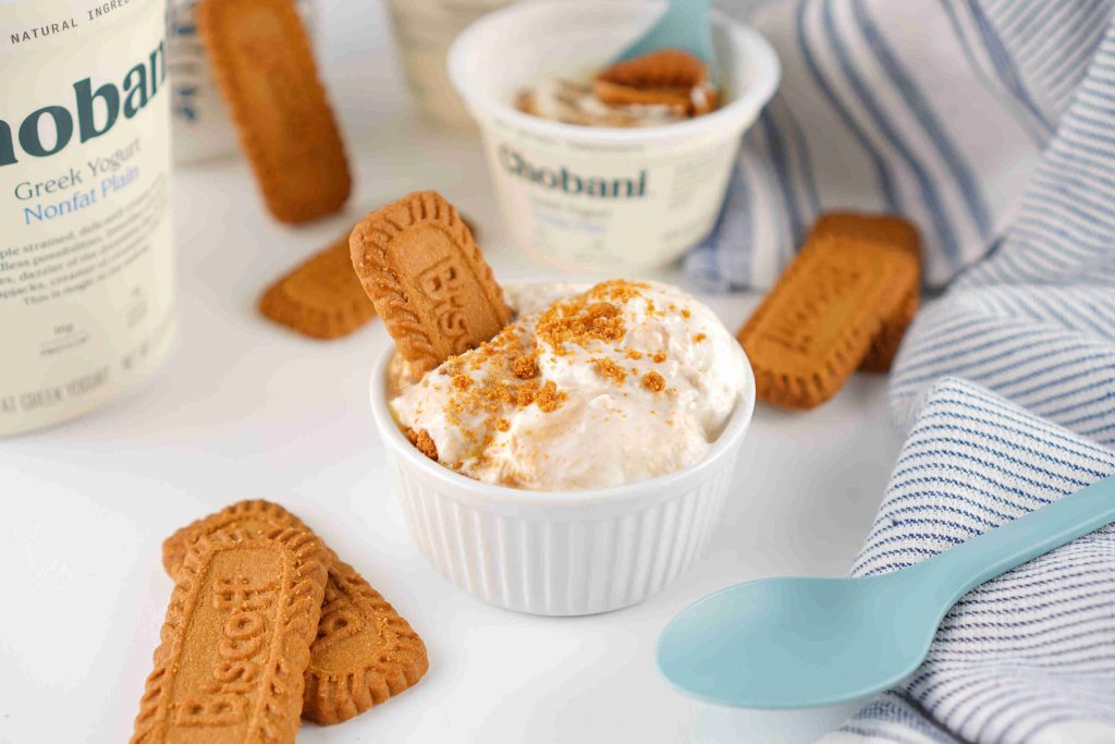 A small white bowl of creamy ice cream topped with crushed Biscoff cookies and a whole cookie—perfect for any Biscoff yogurt recipe—surrounded by more cookies, blue spoons, a striped napkin, and Chobani yogurt containers.