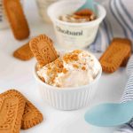 A small white bowl of creamy ice cream topped with crushed Biscoff cookies and a whole cookie—perfect for any Biscoff yogurt recipe—surrounded by more cookies, blue spoons, a striped napkin, and Chobani yogurt containers.