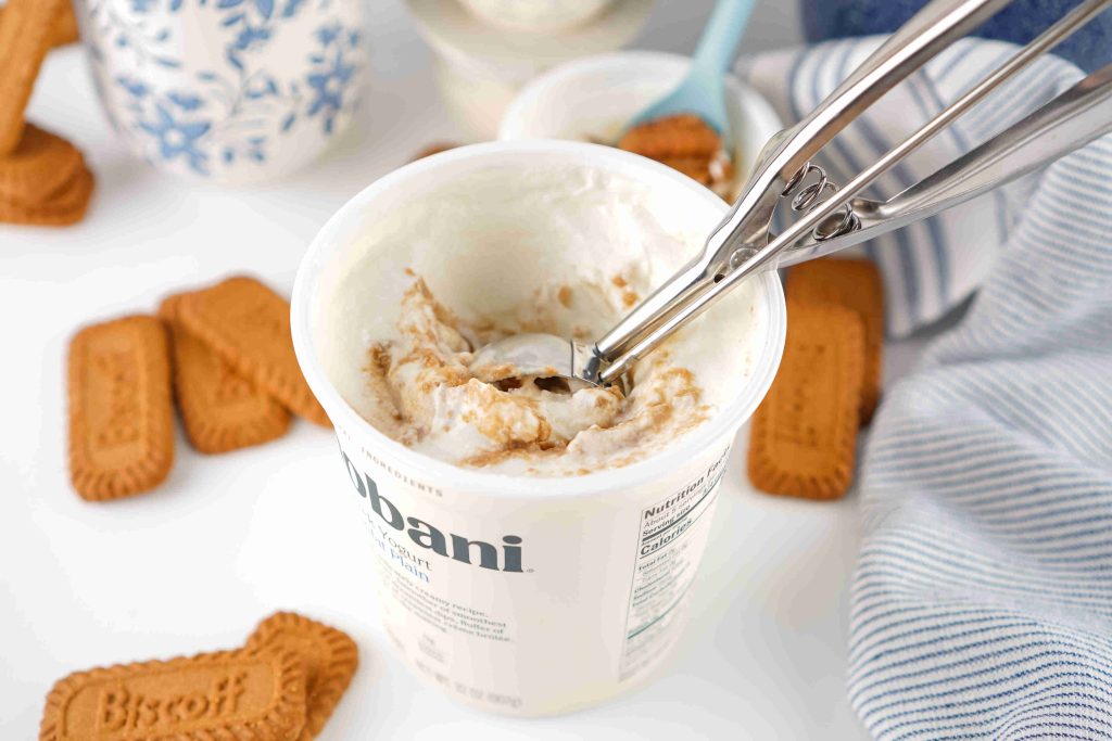 A metal ice cream scoop sits in a container of Chobani with swirls, surrounded by Biscoff cookies and a striped cloth—perfect inspiration for your next Biscoff yogurt recipe.