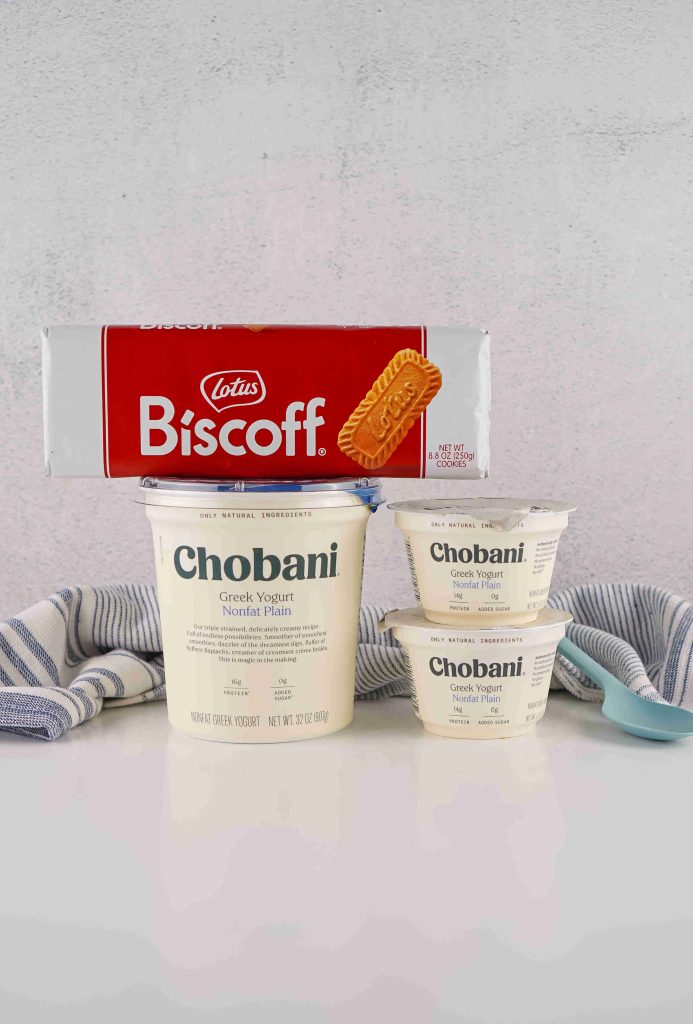 A package of Lotus Biscoff cookies rests atop one large and two small containers of Chobani Greek Yogurt, set on a white surface with a blue-striped towel and spoon—perfect inspiration for your next biscoff yogurt recipe.