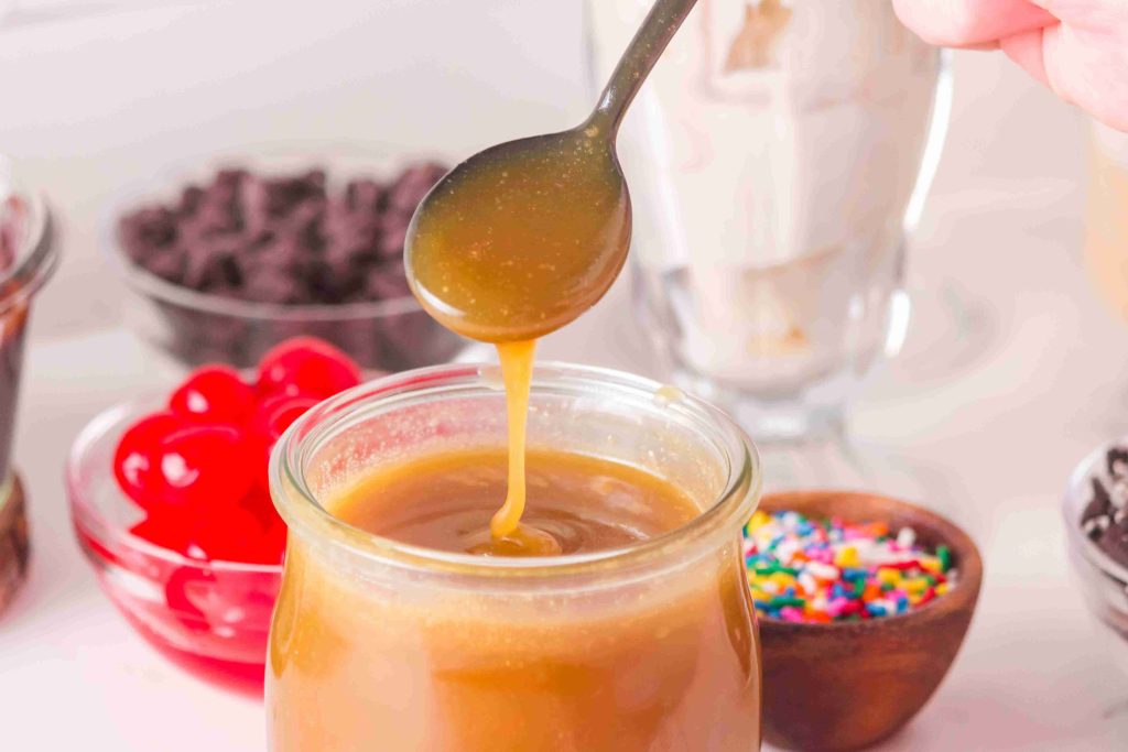 A spoon drizzles caramel sauce into a glass jar, perfect for pairing with your favorite butterscotch sauce recipe. In the background are red cherries, chocolate chips, a milkshake, colorful sprinkles, and other dessert toppings.