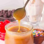 A spoon drizzles caramel sauce into a glass jar, perfect for pairing with your favorite butterscotch sauce recipe. In the background are red cherries, chocolate chips, a milkshake, colorful sprinkles, and other dessert toppings.