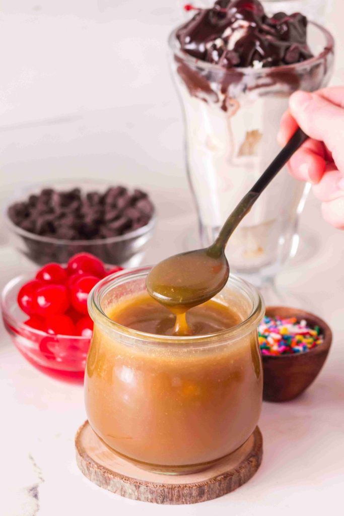 A spoon drizzles caramel sauce from a jar. In the background are a glass of ice cream with chocolate, a bowl of cherries, chocolate chips, and a small dish of rainbow sprinkles—perfect companions if you’re searching for inspiration for your next butterscotch sauce recipe.