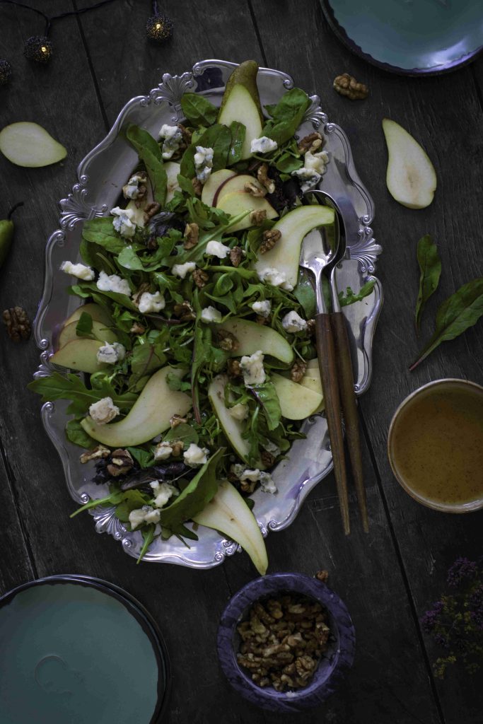 A silver platter holds an Easy Pear and Walnut Salad, featuring a mix of arugula, fresh pear slices, crumbled cheese, and walnuts. Two wooden utensils rest beside it. Surrounding the platter are plates, a bowl of walnuts, salad dressing, and a few salad leaves—an inviting spread perfect for your next Fall Harvest Salad Recipe.