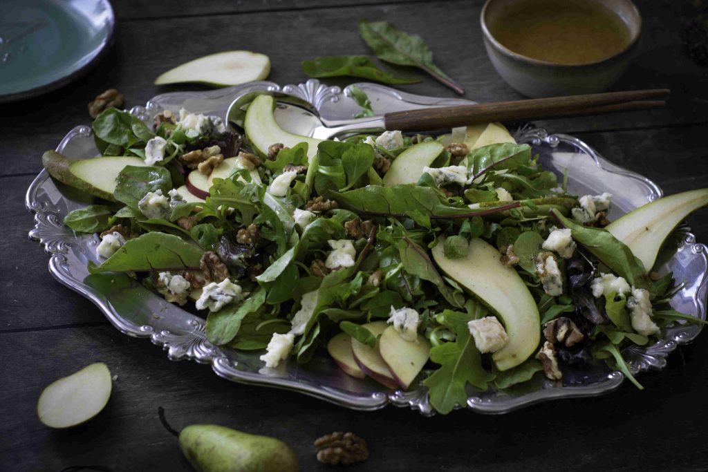 A platter with leafy salad, pear slices, walnuts, and crumbled blue cheese sits on a dark wooden table—an Easy Pear and Walnut Salad complemented by salad dressing, a fork, and scattered pear slices and walnuts.