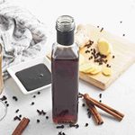 A bottle of gingerbread syrup sits on a white table with a latte, cinnamon sticks, sliced ginger, whole cloves, and a small dish of syrup nearby. A patterned cloth and a wooden board are in the background.
