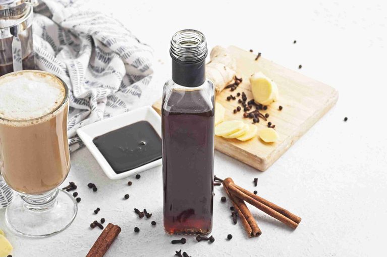A bottle of gingerbread syrup sits on a white table with a latte, cinnamon sticks, sliced ginger, whole cloves, and a small dish of syrup nearby. A patterned cloth and a wooden board are in the background.
