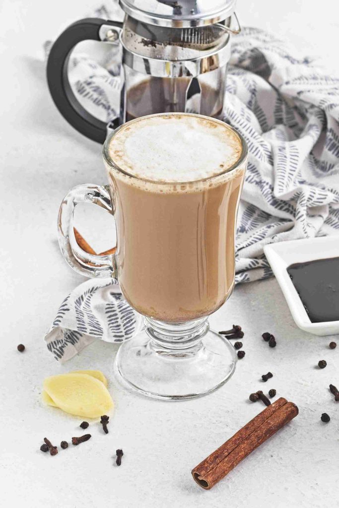 A glass mug of chai latte made with gingerbread syrup sits on a white surface with a cinnamon stick, ginger slices, black pepper, and a French press in the background, next to a patterned cloth.