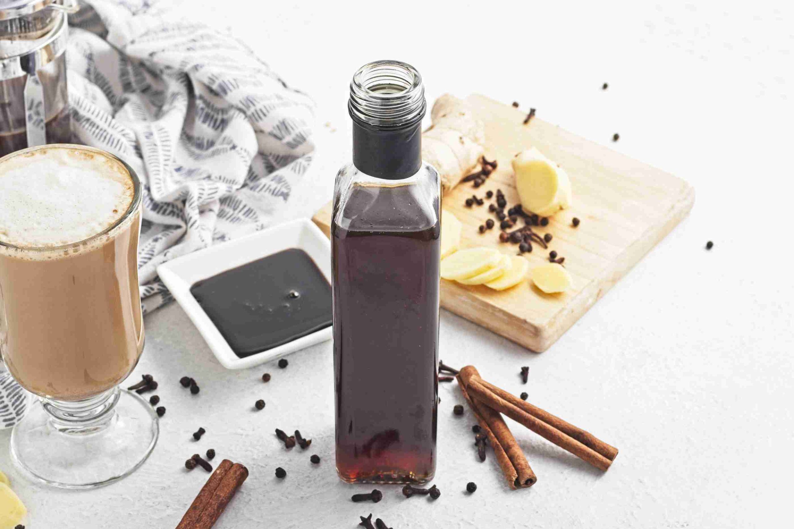 A bottle of gingerbread syrup sits on a white table with a latte, cinnamon sticks, sliced ginger, whole cloves, and a small dish of syrup nearby. A patterned cloth and a wooden board are in the background.
