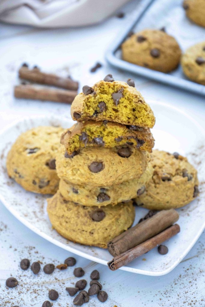 A stack of chocolate chip cookies on a white plate, with two cookies broken open on top. Cinnamon sticks and chocolate chips are scattered around, giving a hint of warmth and spice—perfect if you’re searching for an irresistible pumpkin cookies with chocolate chips treat or a soft pumpkin cookie recipe to enjoy this season.