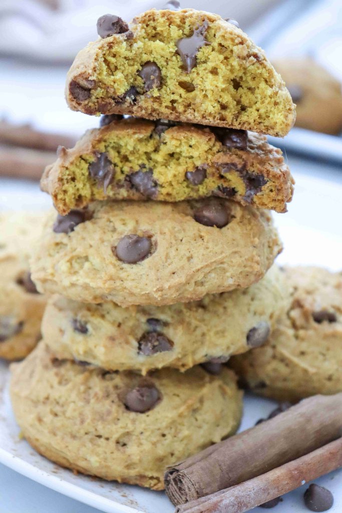 A stack of chocolate chip cookies on a plate, with two broken in half on top, showing gooey chocolate inside. Cinnamon sticks are next to the cookies, evoking the cozy flavors of fall and making you crave a soft pumpkin cookie recipe just as irresistible as these pumpkin cookies with chocolate chips.