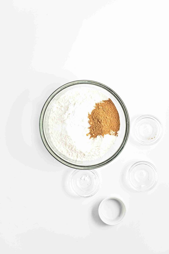 A glass bowl filled with flour and a heap of brown spice sits on a white surface, surrounded by three small empty glass containers—perfect for preparing the dry ingredients for vegan pumpkin chocolate chip cookies.