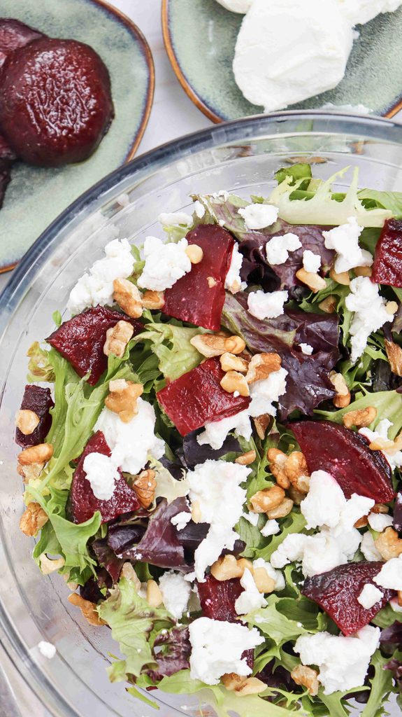A glass bowl of salad with mixed greens, roasted beet slices, crumbled goat cheese, and walnuts makes for a delicious fall salad with beets. Plates nearby hold extra beets and goat cheese.
