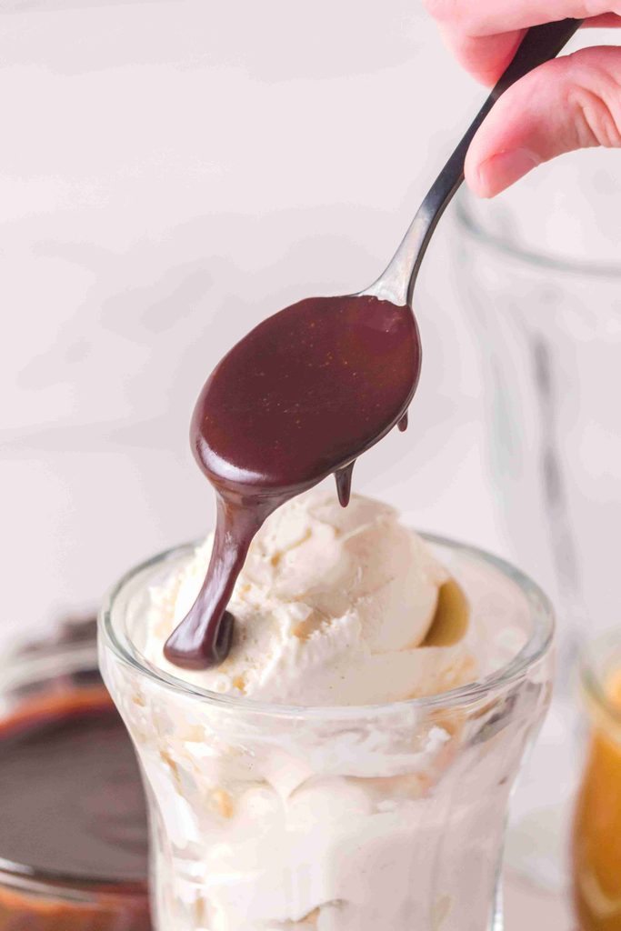 A spoon drizzles rich homemade hot fudge sauce over a glass filled with vanilla ice cream.