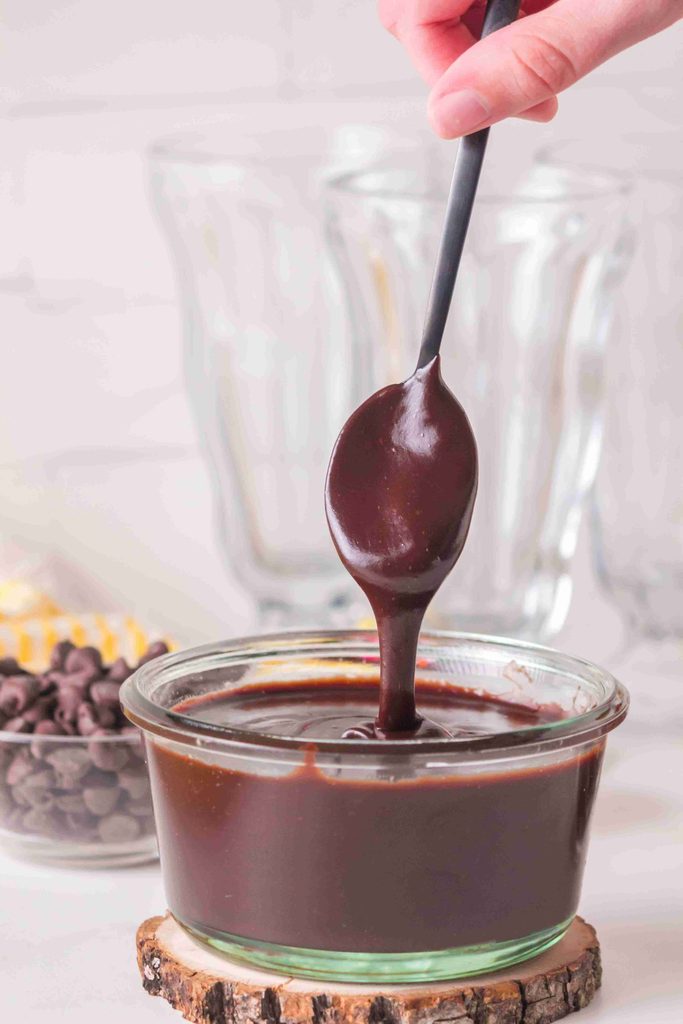 A hand holds a spoon dripping thick, homemade hot fudge sauce over a glass bowl filled with more sauce. Chocolate chips and empty glasses are in the background, highlighting this irresistible hot fudge sauce recipe.