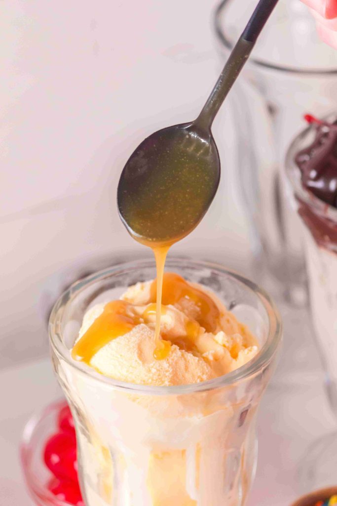 A spoon drizzles caramel sauce over vanilla ice cream in a glass, with whipped cream and cherries visible in the background, reminiscent of a classic butterscotch sauce recipe served at an old-fashioned soda fountain.