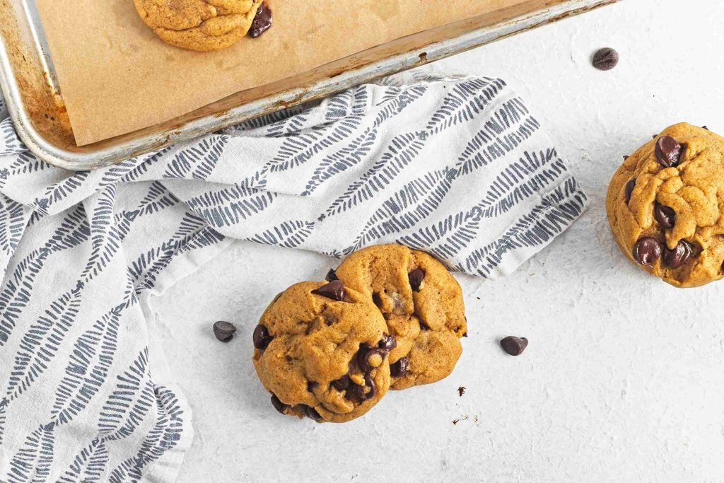 Vegan pumpkin chocolate chip cookies are arranged on a white surface, some resting on a patterned cloth, with a baking tray partially visible in the corner. A few chocolate chips are scattered nearby.