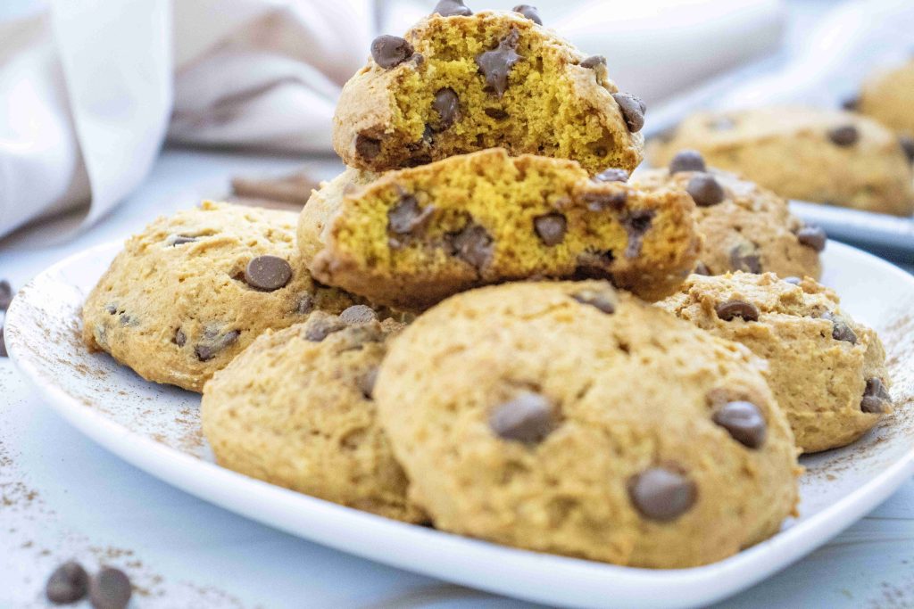 A plate of golden pumpkin cookies with chocolate chips, with one cookie broken in half to reveal a soft, moist interior. These cookies, inspired by a soft pumpkin cookie recipe, are topped with extra chocolate chips and arranged neatly on a white plate.