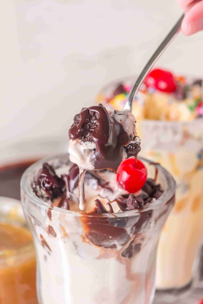 A spoon lifts a bite of ice cream sundae topped with rich homemade hot fudge sauce and a cherry from a glass, with another sundae and a blurred drink in the background.