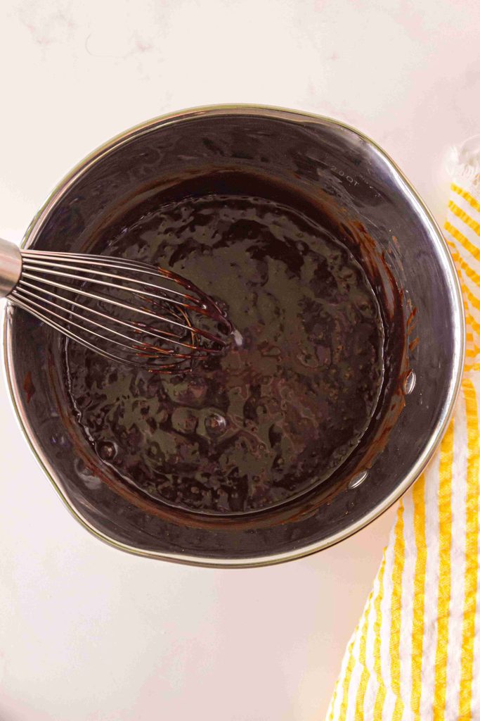 A metal saucepan filled with glossy melted chocolate, perfect for a homemade hot fudge sauce, being mixed with a metal whisk next to a yellow and white striped towel on a white surface.