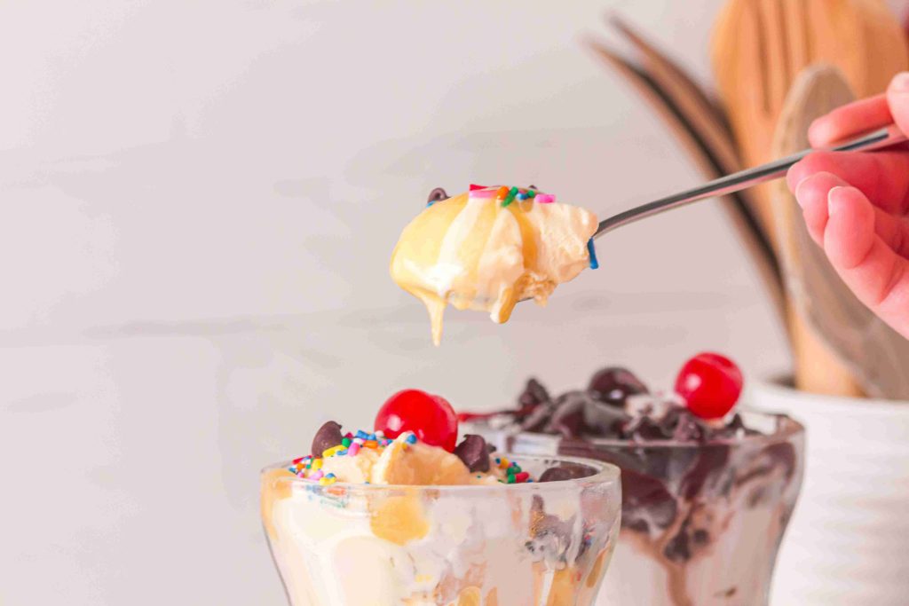 A hand holds a spoonful of ice cream topped with sprinkles, while two dessert glasses below are filled with sundaes, cherries, and chocolate syrup—perfect for drizzling your favorite butterscotch sauce recipe on top.