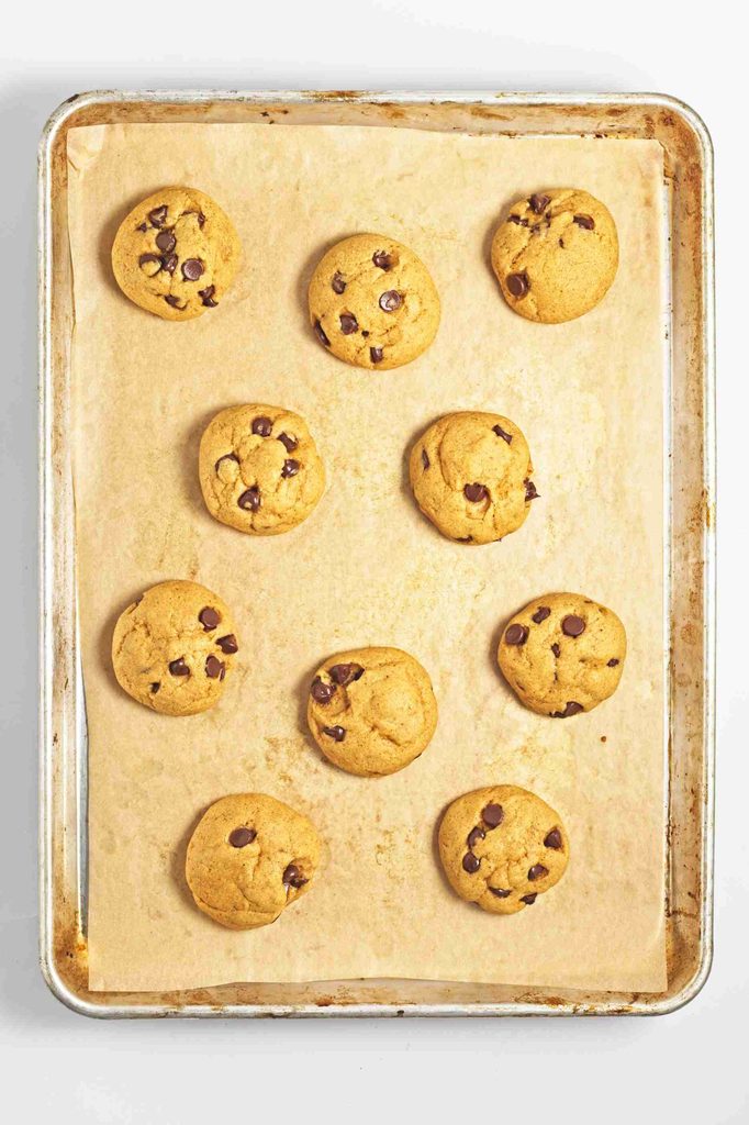 A baking tray lined with parchment paper holds nine freshly baked vegan pumpkin chocolate chip cookies spaced evenly apart.