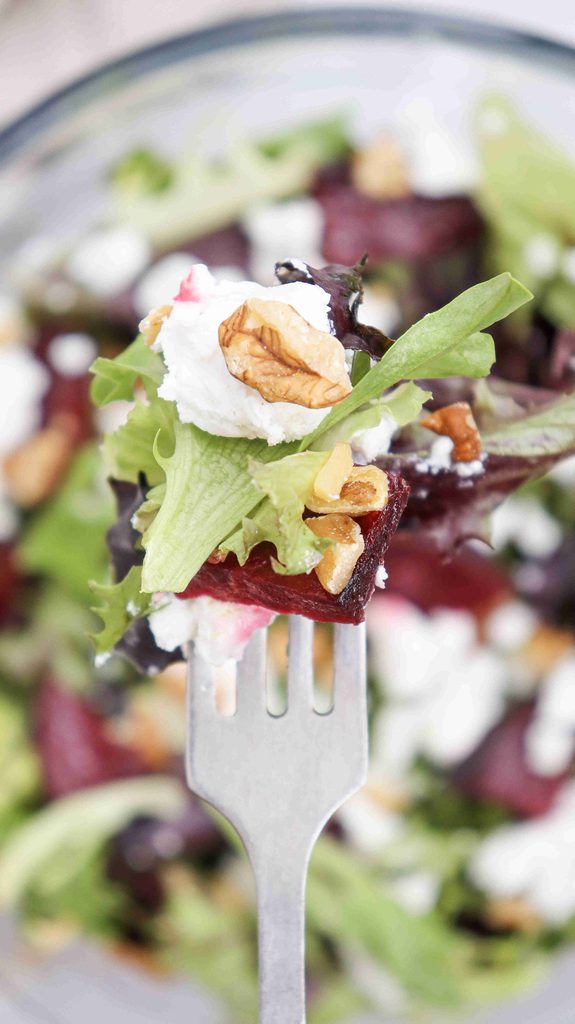 A fork holding mixed greens, beet slices, goat cheese, and a walnut, with a blurred fall salad with beets in the background.