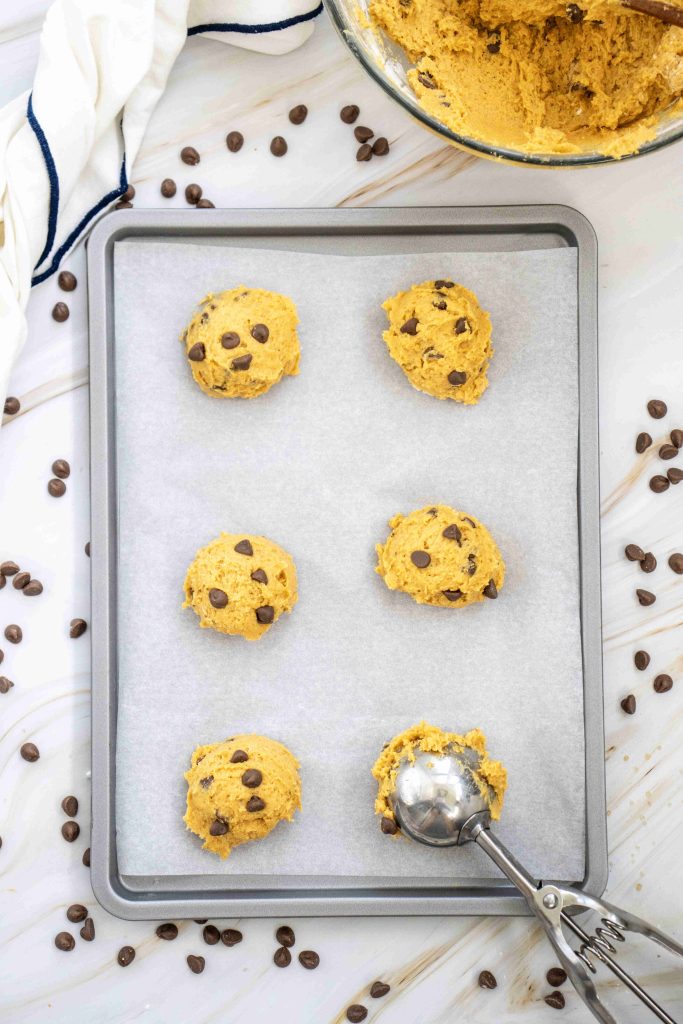 A baking sheet with six scoops of pumpkin cookies with chocolate chips dough on parchment paper, surrounded by scattered chocolate chips and a metal scoop—perfect for trying out your favorite soft pumpkin cookie recipe.