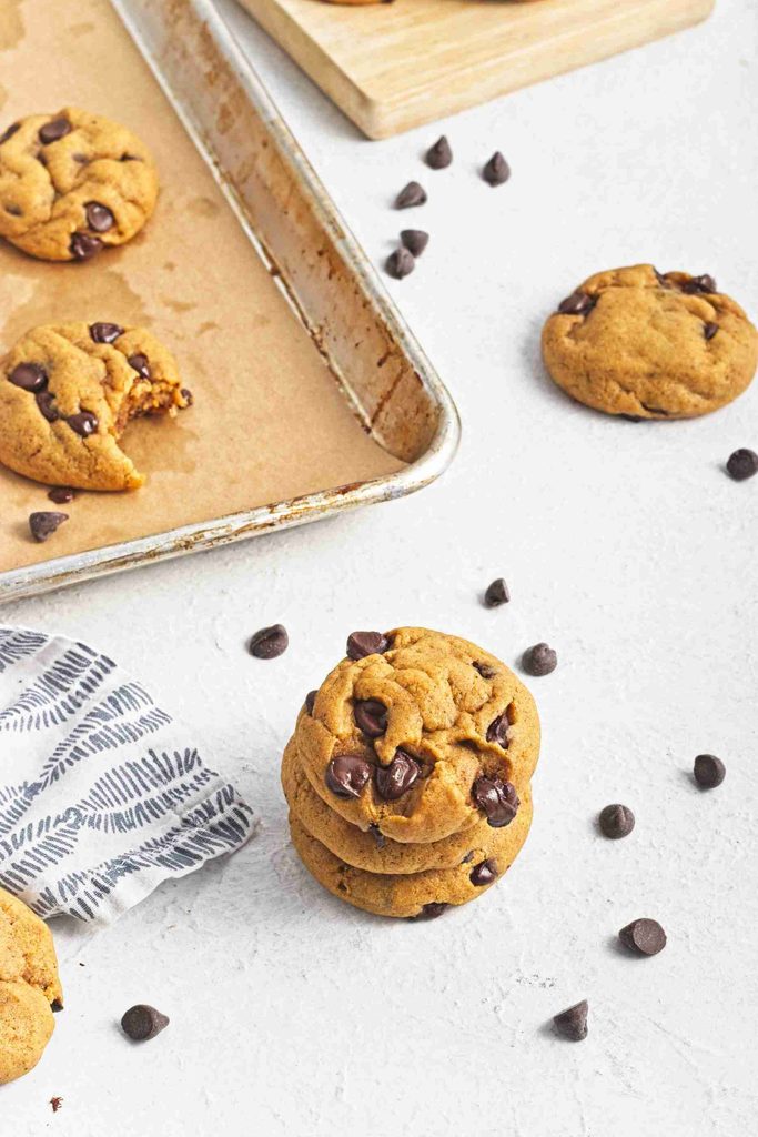 Three vegan pumpkin chocolate chip cookies stacked near a few scattered chocolate chips, with more cookies on a baking sheet lined with parchment paper in the background.